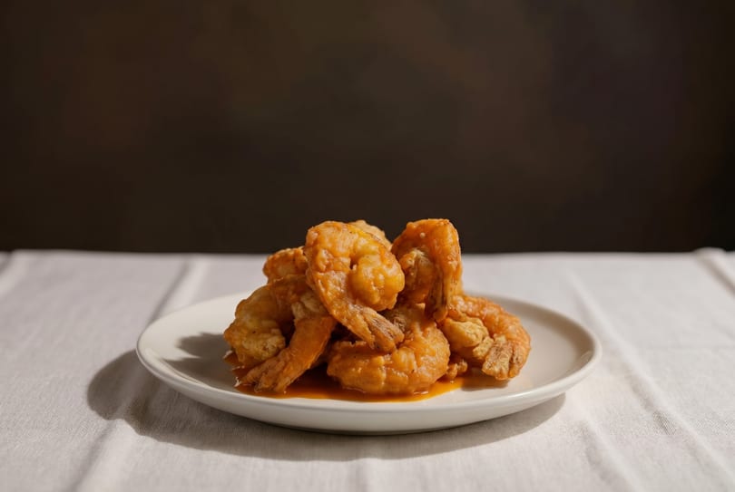 A white plate filled with crispy, golden-brown fried shrimp, glistening with sauce, on a white tablecloth.