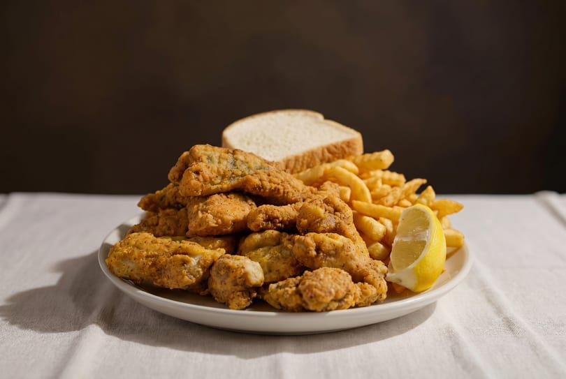 A plate of golden-brown fried fish, french fries, a slice of white bread, and a lemon wedge.
