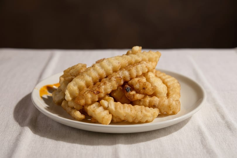 A white plate piled high with golden-brown crinkle-cut fries, with a hint of orange sauce.